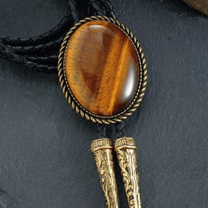 May include: A bolo tie with a large, oval tiger's eye stone set in a gold-toned metal frame. The tie has a black braided leather cord and decorative gold-coloured metal tips with an ornate design. The stone is a warm brown colour.