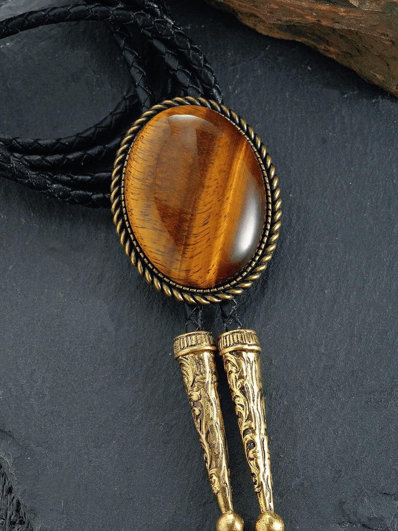 May include: A bolo tie with a large, oval tiger's eye stone set in a gold-toned metal frame. The tie has a black braided leather cord and decorative gold-coloured metal tips with an ornate design. The stone is a warm brown colour.