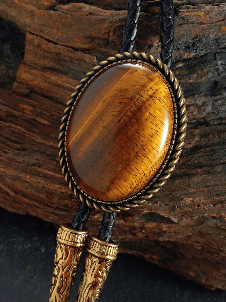 May include: Oval bolo tie featuring a tiger's eye stone. The stone is set in a bronze-coloured metal frame with a rope-like border. The black leather cord has decorative gold-coloured metal tips. A classic accessory.