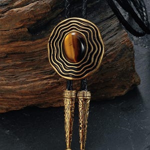 May include: A bolo tie with a gold-colored, layered circular pendant featuring a brown oval stone. The pendant is attached to a black braided leather cord. The tie has two gold-colored decorative tips.