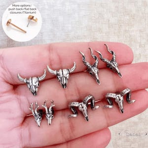 May include: Silver-toned animal skull stud earrings. The earrings feature various designs, including bull, deer, and ram skulls. Push back/flat back closures (Titanium) are shown in the upper left corner. The earrings are displayed on a hand.
