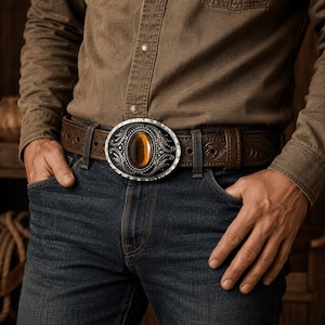 May include: A brown leather belt featuring an oval-shaped silver-tone metal buckle with intricate scrollwork engravings and a central, smooth, polished amber-colored gemstone inlay. The belt strap has a dark brown finish with embossed patterns.