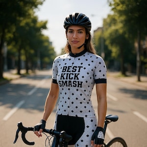 Op de afbeelding: Een vrouw in een wit en zwart fietsjersey met de tekst "BEST KICK SMASH" en zwarte fietsbroek staat met een zwarte racefiets. Ze draagt een zwarte helm en zwarte handschoenen.