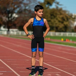 May include: A young person wearing a black and blue athletic suit with a blue and purple camouflage pattern on the legs. The person is standing on a red running track.