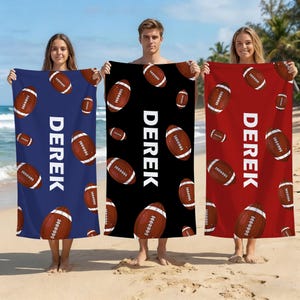 Puede incluir: Tres toallas de playa en azul, negro y rojo, cada una con un patrón repetido de balones de fútbol marrones con cordones blancos. La palabra "DEREK" está impresa verticalmente en blanco en cada toalla. Las toallas son sostenidas por tres personas en una playa de arena.