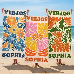 Puede incluir: Tres toallas de playa con diseños florales y el nombre "SOPHIA" en negrita. Las toallas presentan colores vibrantes como azul, naranja, rosa y verde, sobre un fondo crema. Las toallas se sostienen en una playa arenosa.