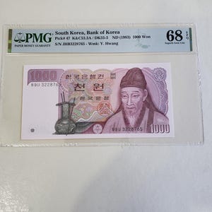 May include: A South Korean 1000 Won banknote from 1983, graded 68 by PMG. The note features a portrait of a man in traditional attire, with Korean text and a vase design. The banknote is encased in a protective holder.