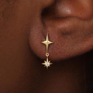 May include: A gold stud earring featuring a four-pointed star design at the lobe, with a small dangling charm shaped like a multi-pointed star that contains a single clear, round gemstone at its center, secured by a thin gold link chain.