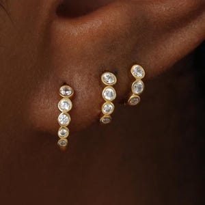 May include: Three gold hoop earrings worn in a single earlobe. Each hoop features a series of small, round, clear stones set in a bezel style. The earrings vary in size, with the largest hoop positioned at the bottom and the smallest at the top.