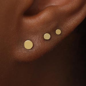May include: Three small, round, flat gold stud earrings are arranged in a row along the earlobe. Each earring features a smooth, polished surface and a simple circular shape, with the sizes graduating from largest to smallest in diameter.