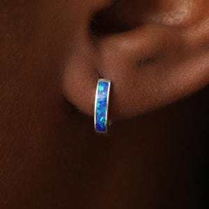 May include: A small silver hoop earring featuring a narrow inlay of iridescent blue opal. The earring is circular in shape with a polished metallic finish, showcasing a vibrant, multi-faceted blue pattern embedded along the outer edge of the band.