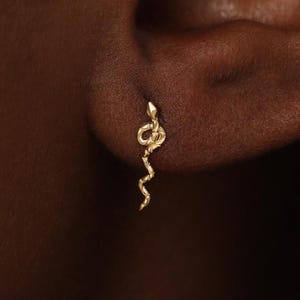 Peut inclure: Une petite boucle d'oreille à clou en or en forme de serpent enroulé. La boucle d'oreille présente une surface texturée et un corps fin et sinueux qui s'étend vers le bas depuis le lobe de l'oreille, avec une tête détaillée et une queue courbée.