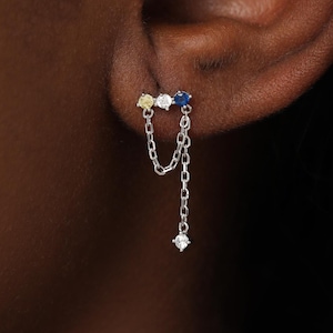 May include: A silver-toned earring featuring three small, multi-colored gemstones in yellow, white, and blue, set in a horizontal row. Two delicate silver chains hang from the setting, with one chain ending in a single, small, clear gemstone.