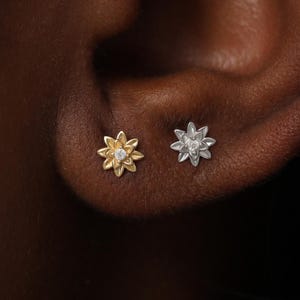 May include: Two small stud earrings shaped like multi-petaled flowers. One earring is gold-toned with a central clear stone, and the other is silver-toned with a central clear stone. Both earrings feature textured, layered petals.