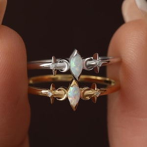 May include: Two rings stacked vertically, one silver-toned and one gold-toned. Each ring features a central marquise-cut iridescent opal stone flanked by small crescent moon shapes and tiny embedded clear stones on a thin, polished metal band.