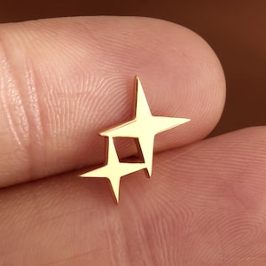 May include: A small gold-toned stud earring featuring a design of two four-pointed stars connected at their tips. The metal has a smooth, polished finish with sharp, angular points extending from the center of each star shape in a symmetrical pattern.