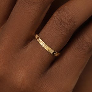 May include: A thin gold band ring featuring a delicate, repeating floral or botanical pattern engraved along the entire outer surface. The metal has a polished finish that reflects light, highlighting the intricate details of the small, etched motifs.