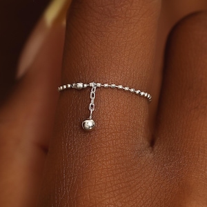 Op de afbeelding: Een zilverkleurige ring met een delicate kralenband. Een korte, dunne schakelketting hangt aan het midden van de band en eindigt in een kleine, gepolijste zilverkleurige bolvormige bedel die tegen de vinger rust.