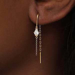 May include: A gold threader earring featuring a delicate chain, a small pear-shaped clear gemstone, and a thin straight metal bar at the end. The earring is inserted through the earlobe, with the chain and bar hanging down from the back.
