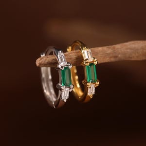 May include: Two small hoop earrings, one silver-toned and one gold-toned, each featuring a central rectangular green gemstone flanked by two smaller clear baguette-cut stones. The earrings are displayed side-by-side on a thin wooden branch.
