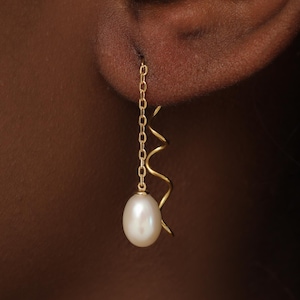 May include: A gold earring featuring a delicate chain and a wavy gold wire element, finished with a single white oval pearl drop. The earring hangs vertically from the earlobe, showcasing a combination of metallic gold and a smooth pearl.