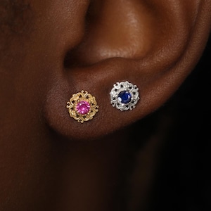 May include: Two stud earrings featuring intricate filigree metalwork. One earring is gold-toned with a round pink center stone, and the other is silver-toned with a round blue center stone. Both earrings have a circular, ornate, scalloped edge.
