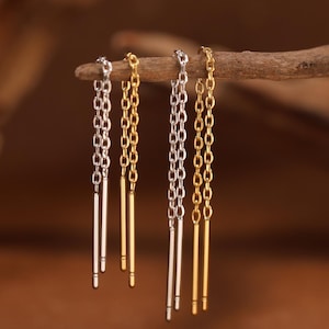 May include: Two pairs of threader earrings, one pair in silver and one pair in gold. Each earring consists of a delicate chain connected to a thin, straight metal bar at the end. The earrings are suspended from a piece of textured, weathered wood.