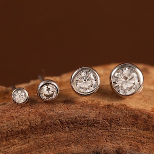 May include: Four silver-toned stud earrings featuring round, clear, faceted stones set in a bezel setting. The earrings are arranged in a row from smallest to largest, showcasing a metallic finish and reflective, sparkling surfaces.