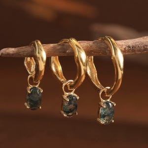 May include: Three gold hoop earrings with an irregular, textured surface. Each hoop features a small, oval-shaped moss agate gemstone charm dangling from the bottom. The gemstones display deep green and translucent inclusions within the stone.