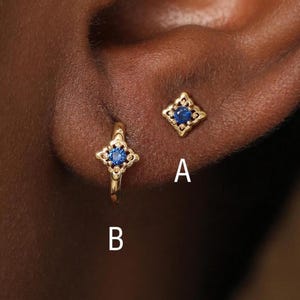 Puede incluir: Dos pendientes de oro con una piedra preciosa azul engastada en un marco en forma de estrella de cuatro puntas. El pendiente A es un botón y el pendiente B es un pequeño aro. Ambas piezas están marcadas con las letras A y B.