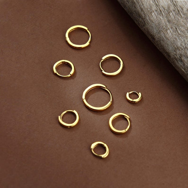 May include: A collection of eight gold-toned hoop earrings of varying sizes arranged in a scattered pattern. Each earring features a simple, circular design with a smooth, polished finish and a hinged closure mechanism for secure fastening.