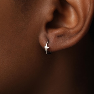May include: A small silver hoop earring featuring a four-pointed star charm. The star has sharp, elongated points and a polished metallic finish. The earring is secured through the earlobe, showcasing a simple and clean design with a reflective surface.