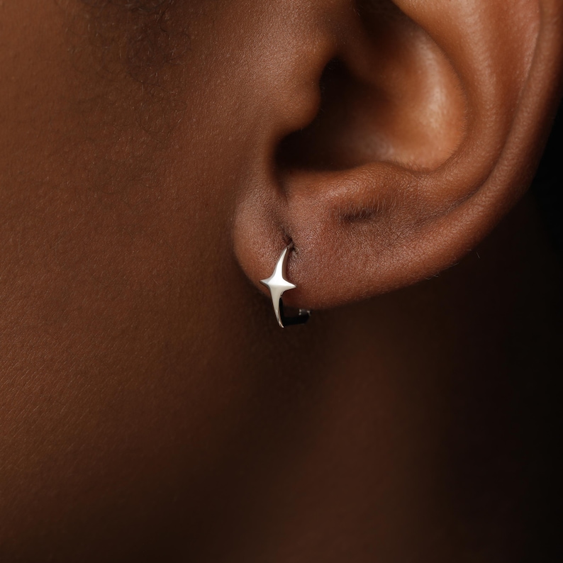 May include: A small silver hoop earring featuring a four-pointed star charm. The star has sharp, elongated points and a polished metallic finish. The earring is secured through the earlobe, showcasing a simple and clean design with a reflective surface.