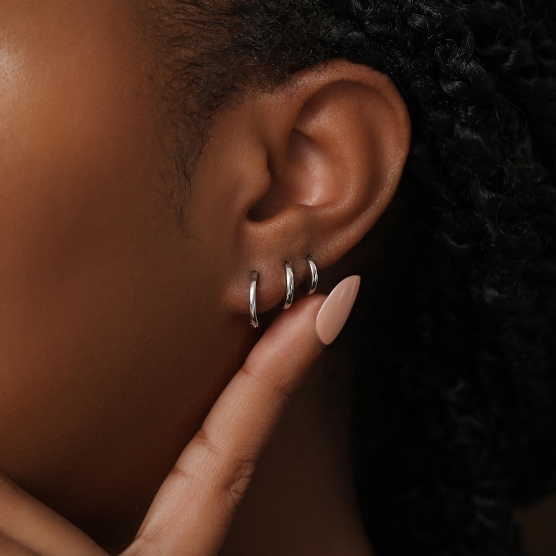 May include: Three small, polished silver hoop earrings arranged in a row along the earlobe. The hoops are circular, metallic, and have a smooth, reflective surface. Each earring is a simple, thin band design, creating a uniform look across the ear.