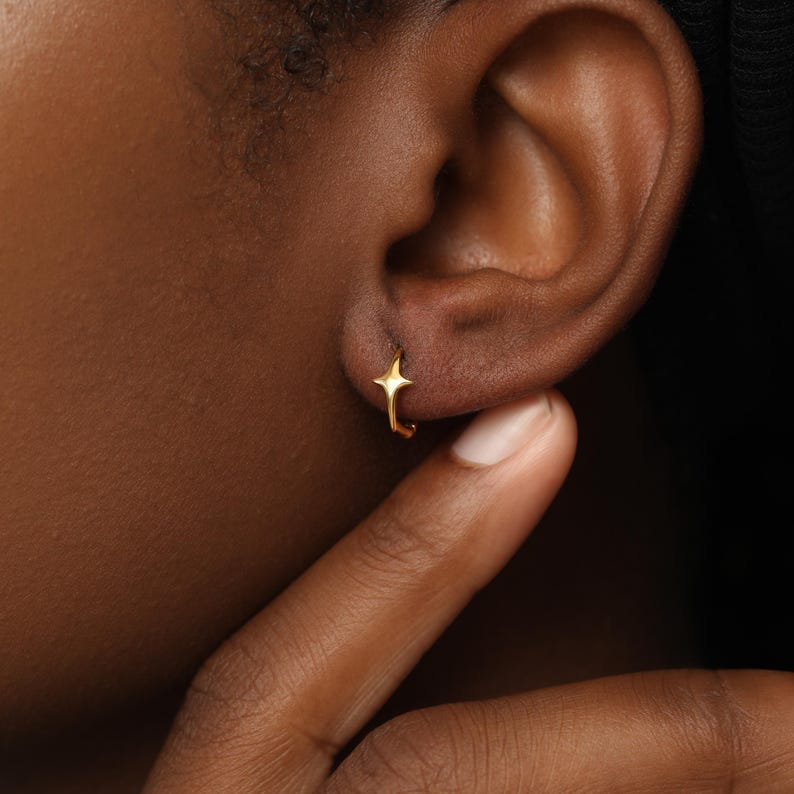 May include: A small gold hoop earring featuring a four-pointed star charm. The earring is circular in shape with a polished metallic finish, and the star element is centered on the front of the hoop, creating a distinct, symmetrical design.