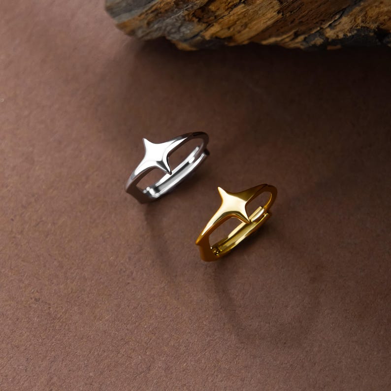 May include: Two rings, one in a silver-toned metal and one in a gold-toned metal, each featuring a prominent four-pointed star design at the center of the band. The rings have a smooth, polished finish and a simple, open-ended band structure.