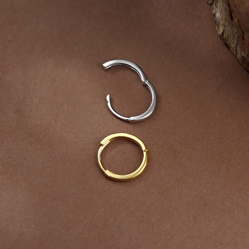 May include: Two small hoop earrings, one in a polished silver tone and one in a polished gold tone. Each earring features a hinged closure mechanism, allowing the hoop to open and close securely. The earrings are circular in shape and smooth.