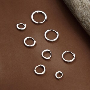 May include: A collection of seven silver-toned hoop earrings of varying sizes arranged in a scattered pattern. Each earring features a simple, circular design with a hinged closure mechanism, showcasing a polished metallic finish throughout the set.