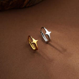 May include: Two small hoop earrings, one in a polished gold tone and one in a polished silver tone. Each earring features a prominent four-pointed star design centered on the outer edge of the hoop, creating a distinct geometric aesthetic.