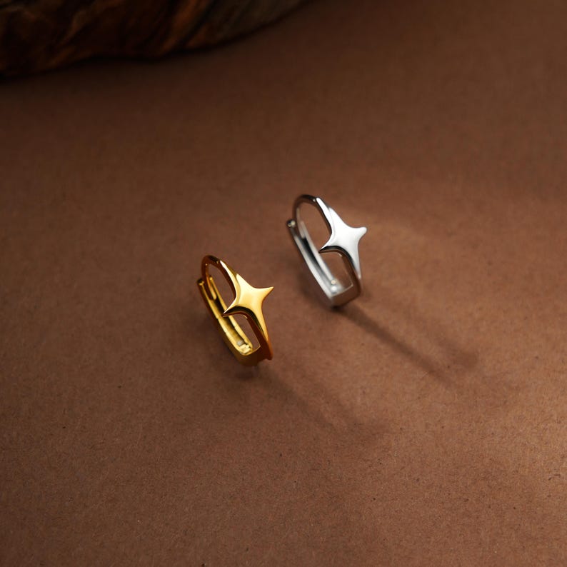May include: Two small hoop earrings, one in a polished gold tone and one in a polished silver tone. Each earring features a prominent four-pointed star design centered on the outer edge of the hoop, creating a distinct geometric aesthetic.