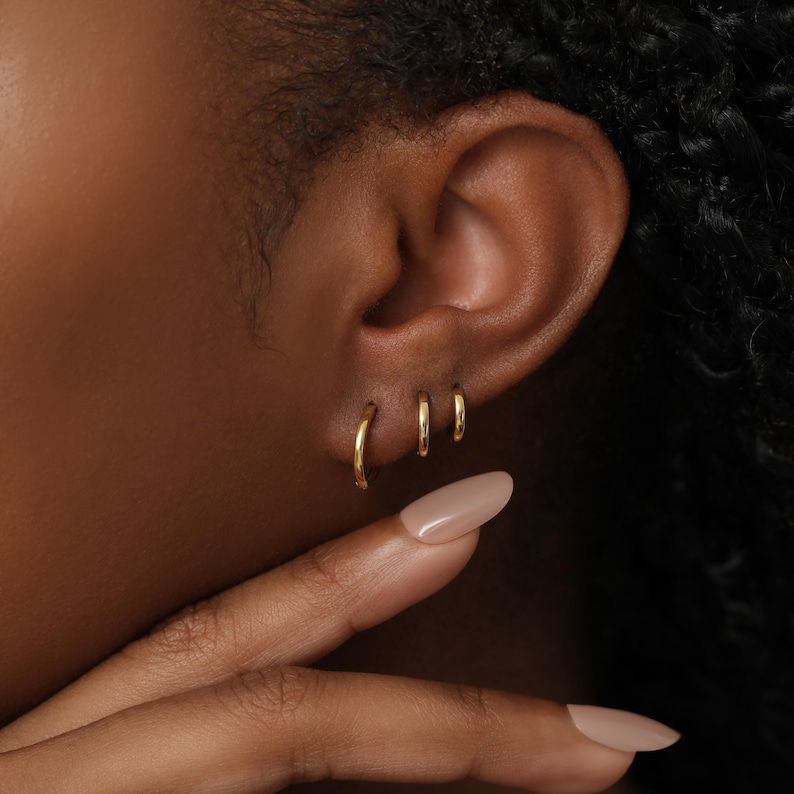 May include: Three small, polished gold hoop earrings of varying sizes arranged in a row along the earlobe. The hoops feature a smooth, rounded metal surface and a simple circular design, creating a clean and understated look for the ear.