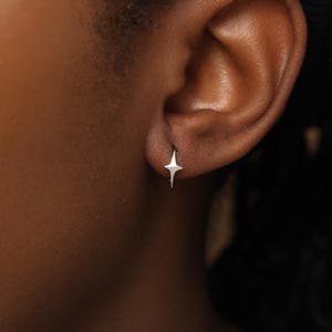 May include: A small, silver-toned earring shaped like a four-pointed star. The earring features a polished, metallic finish with sharp, elongated points extending from the center, securely fastened to the earlobe in a simple, minimalist design.