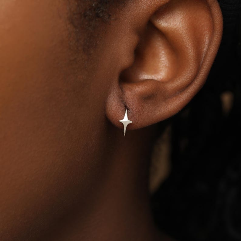 May include: A small, silver-toned earring shaped like a four-pointed star. The earring features a polished, metallic finish with sharp, elongated points extending from the center, securely fastened to the earlobe in a simple, minimalist design.