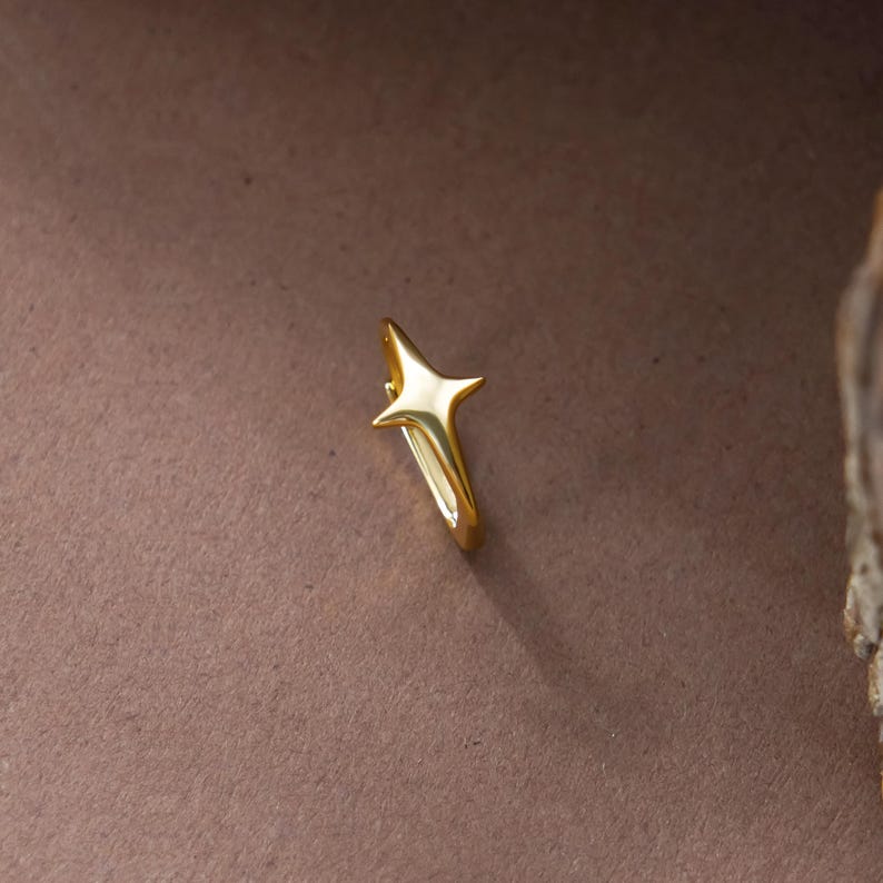 May include: A gold-toned hoop earring featuring a four-pointed star charm. The star has a smooth, polished surface with elongated, tapered points. The hoop is circular and connects to the base of the star, creating a cohesive metallic piece.