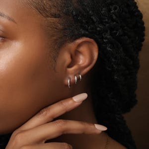 May include: Three small, polished silver hoop earrings arranged in a row along the earlobe. Each hoop features a simple, circular design with a smooth, reflective metallic finish. The earrings are worn in three consecutive piercings on the ear.