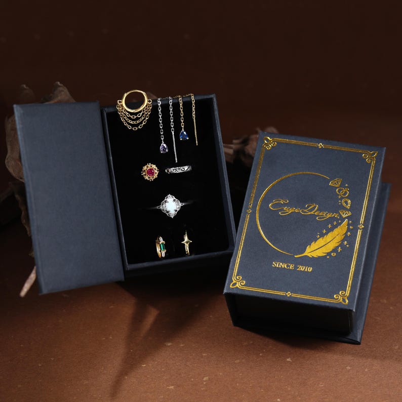May include: A black jewelry box with gold foil text reading EugeDesign SINCE 2010 and a feather graphic. Inside, a black velvet insert holds a gold hoop earring with chains, two drop earrings with blue stones, and four rings featuring red, white, and green stones.
