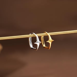 May include: Two small hoop earrings, one silver-toned and one gold-toned, each featuring a four-pointed star design on the outer edge. The earrings are suspended on a thin, horizontal gold-colored rod, showcasing their polished metallic finish.