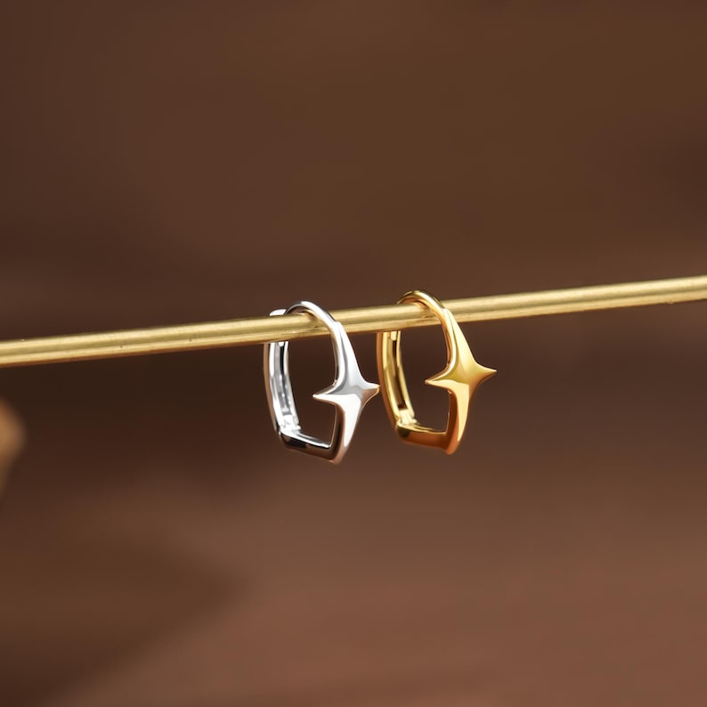 May include: Two small hoop earrings, one silver-toned and one gold-toned, each featuring a four-pointed star design on the outer edge. The earrings are suspended on a thin, horizontal gold-colored rod, showcasing their polished metallic finish.