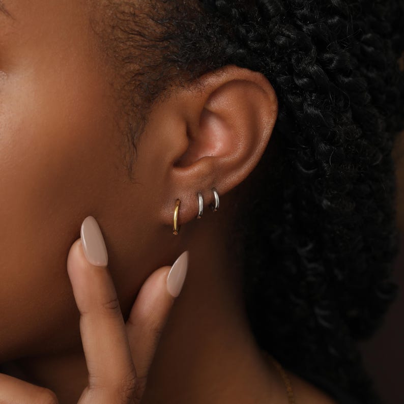 May include: Three small hoop earrings worn in a row on an earlobe. The earring closest to the face is gold-toned, while the two smaller hoops positioned behind it are silver-toned. Each hoop features a smooth, polished, and circular metal design.