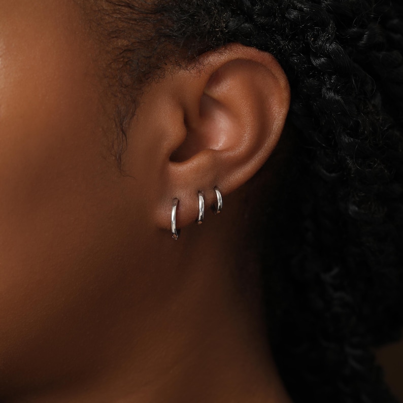May include: Three small, polished silver-tone hoop earrings of varying sizes arranged in a row along the earlobe. The hoops have a smooth, circular profile and a reflective metallic finish, creating a simple and clean look for the ear.
